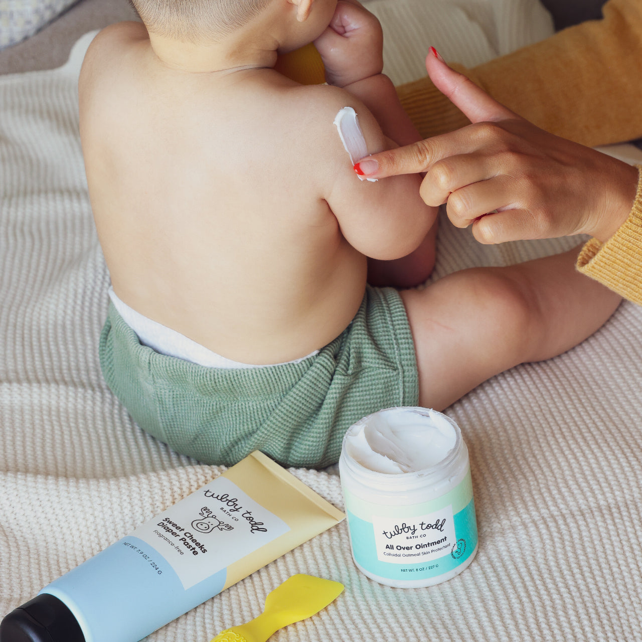 Mama applying cream to a baby's skin with a jar of AOO and Sweet Cheeks Diaper Paste visible on a blanket.